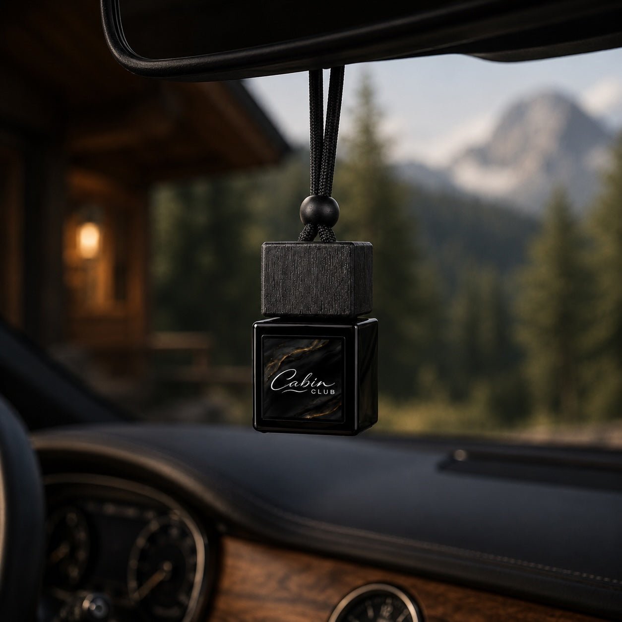 Cabin Club Alpine Cabin car fragrance diffuser hanging from a rear-view mirror with a mountain lodge behind it
