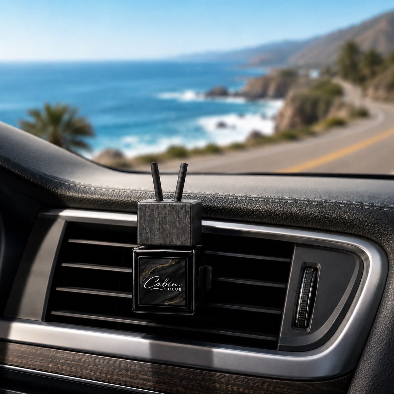 Cabin Club Coastal Drive car fragrance diffuser clipped to an air vent beside a scenic ocean road