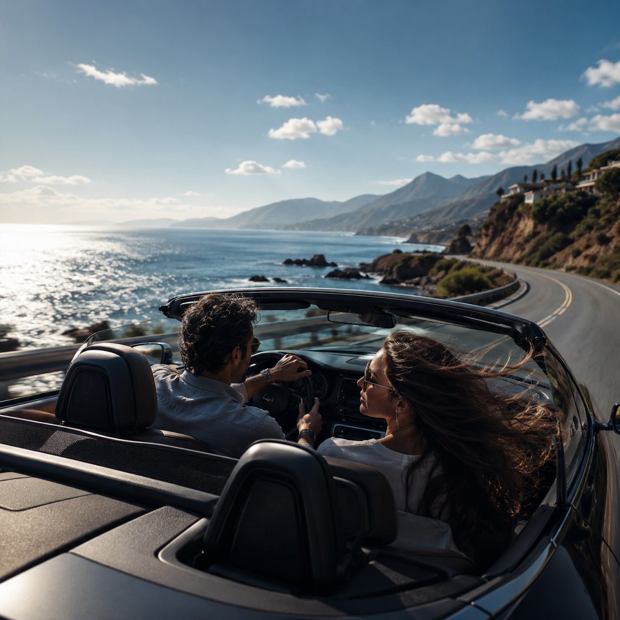 Couple driving a black convertible along a sunlit coastal road beside the ocean and mountains