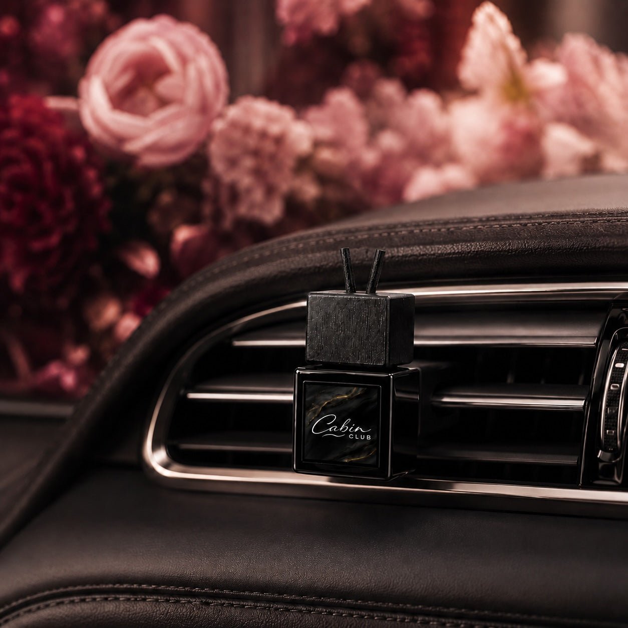 Cabin Club Velvet Bloom car fragrance diffuser clipped to an air vent with pink and burgundy flowers behind it